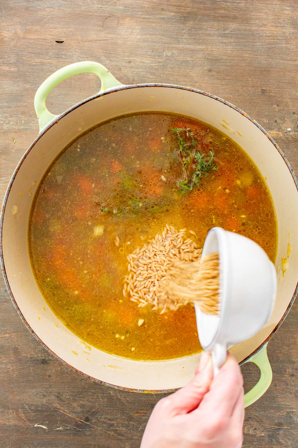 A hand pours dry orzo into the simmering broth and vegetables in a Dutch oven.