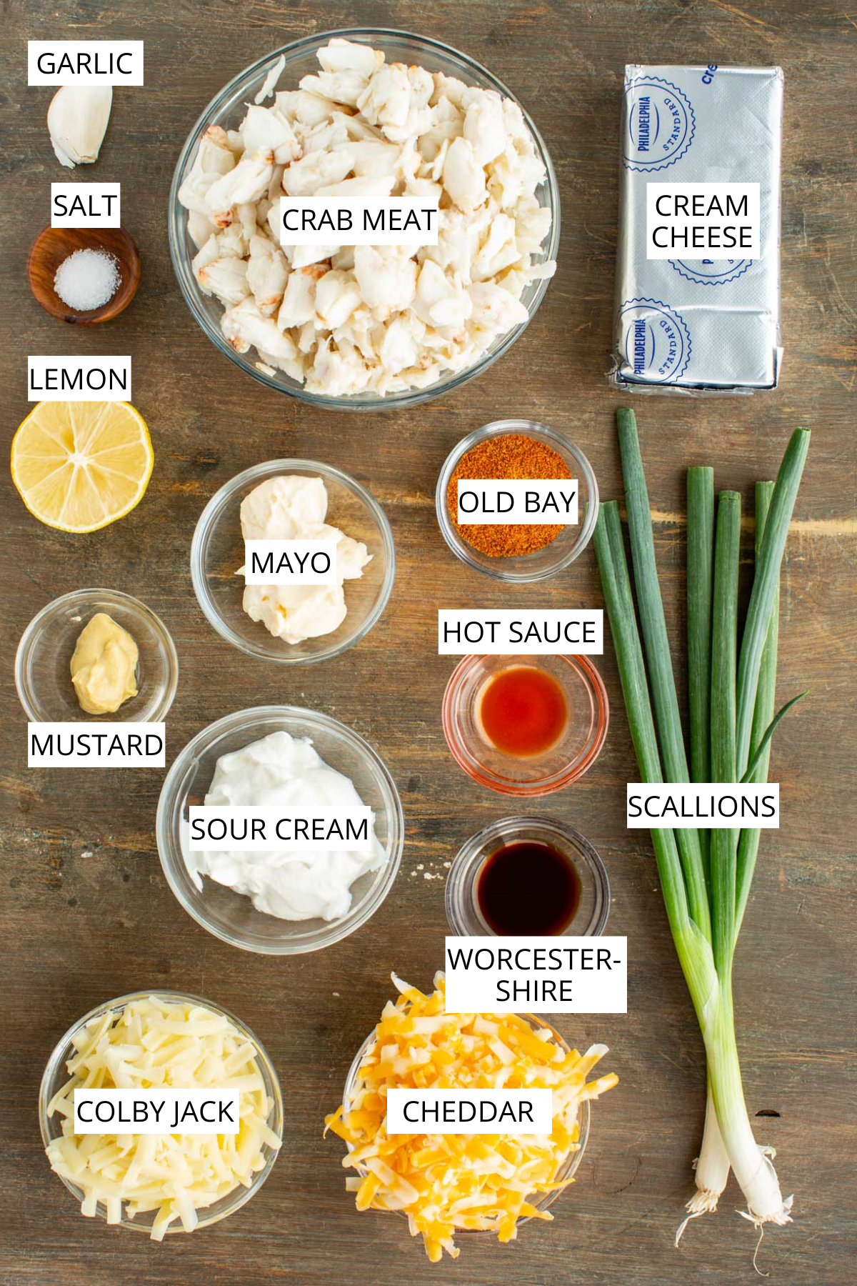 All the ingredients for Maryland crab dip arranged on a wooden surface, including lump crabmeat, cream cheese, sour cream, mayonnaise, cheddar, colby jack, scallions, lemon, garlic, mustard, Old Bay and hot sauce.