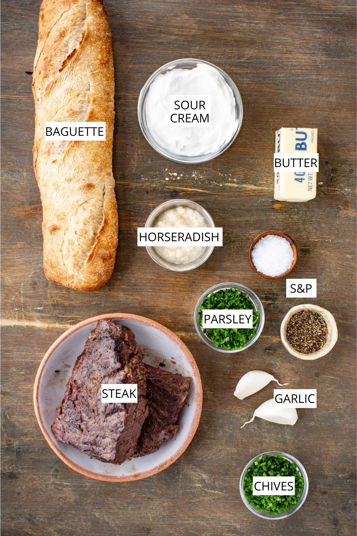 All ingredients for steak crostini laid out on a wooden surface including baguette, sour cream, horseradish, butter, parsley, garlic, chives, salt, pepper and cooked steak.