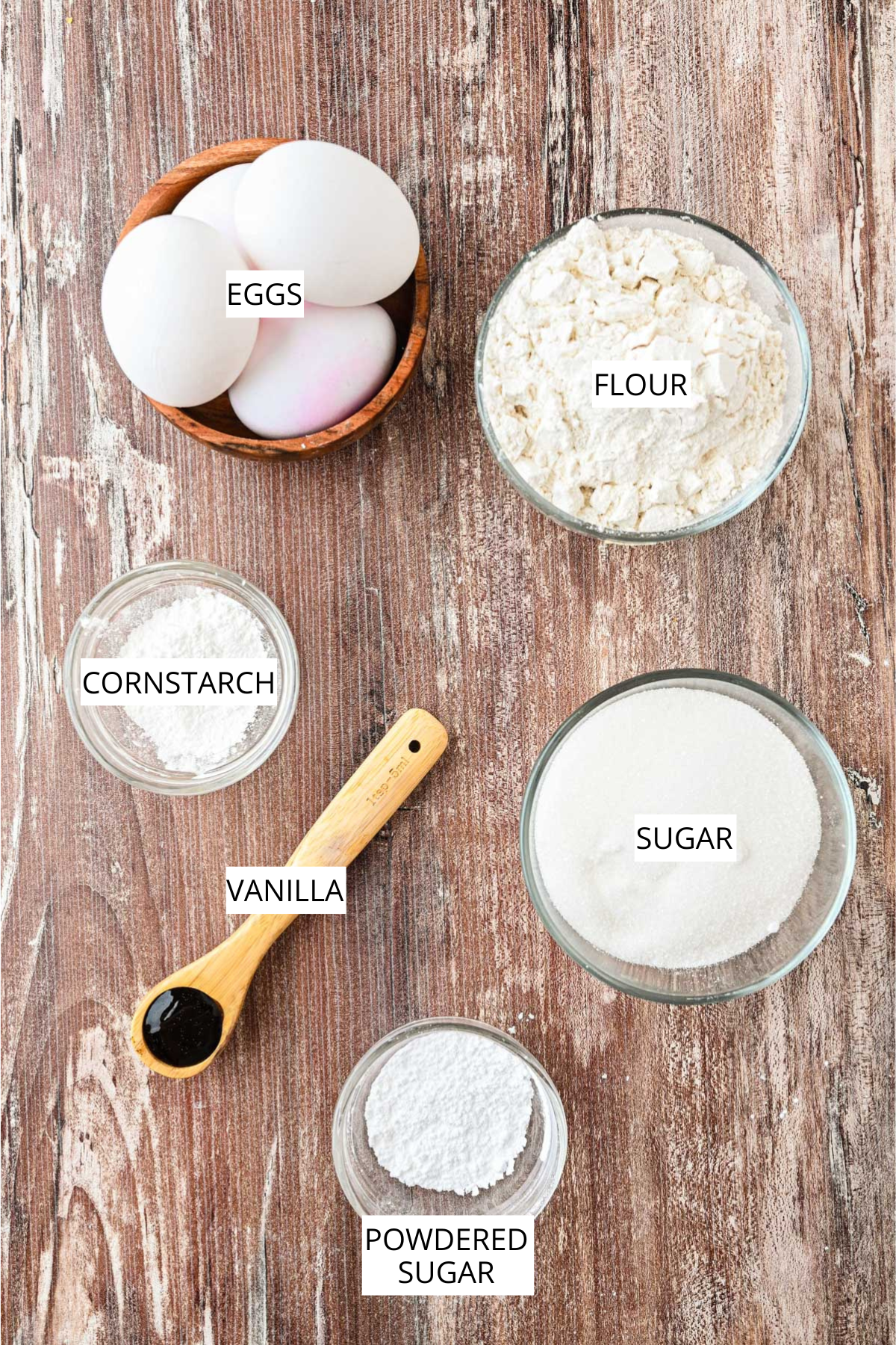 Bowls of flour, sugar, eggs, cornstarch, vanilla, and powdered sugar arranged on a rustic wood surface, each labeled.