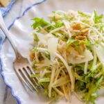 Thinly shaved celery root and pear tossed with frisee, walnuts and manchego on a white plate with a fork.