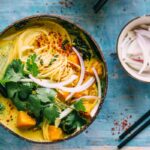 An overhead view of coconut curry noodle soup garnished with cilantro, sliced red onion, chili flakes, and lime, set on a blue surface.