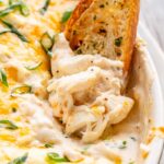 Creamy baked crab dip with large chunks of lump crabmeat being scooped up with a piece of toasted bread, garnished with melted cheese and sliced scallions.