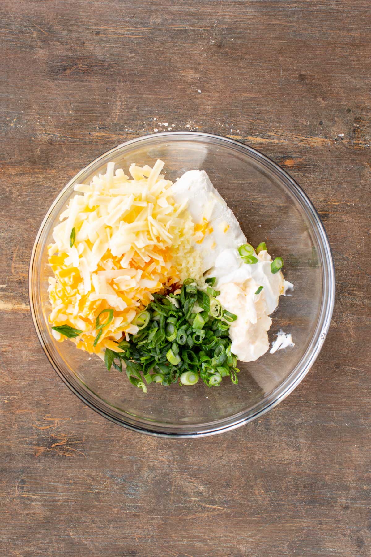 A glass bowl filled with shredded cheese, cream cheese, sour cream, mayonnaise, scallions and garlic sitting on a wooden surface.