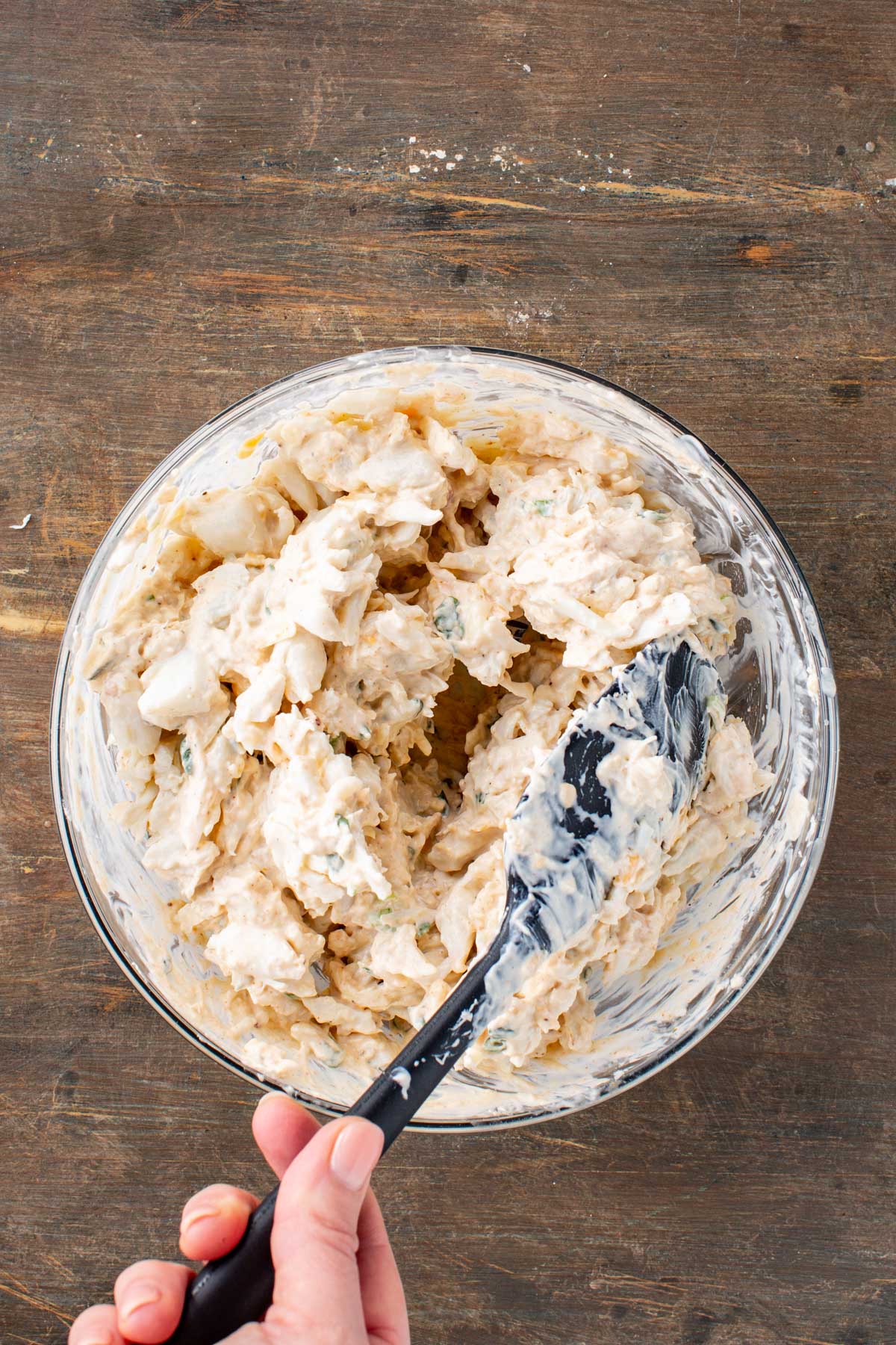 A hand using a spatula to gently fold lump crabmeat into the creamy cheese base.