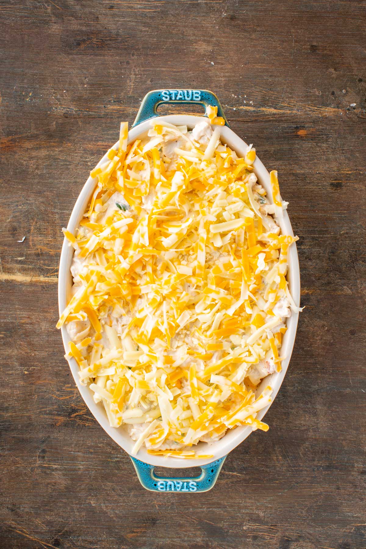 An oval baking dish filled with the crab mixture topped with shredded cheddar and jack cheese.