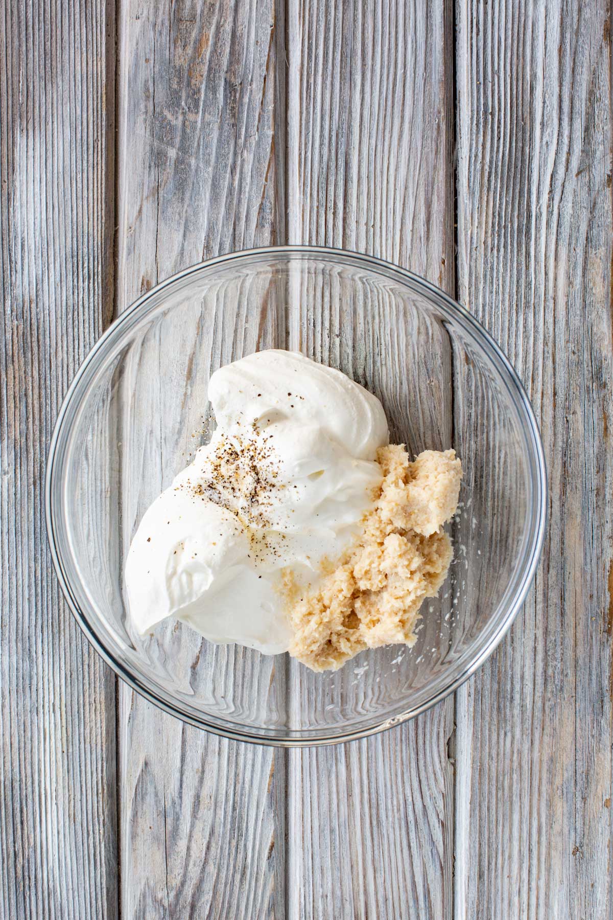 Sour cream and prepared horseradish in a clear glass bowl with cracked black pepper on top, set on a weathered wooden surface.