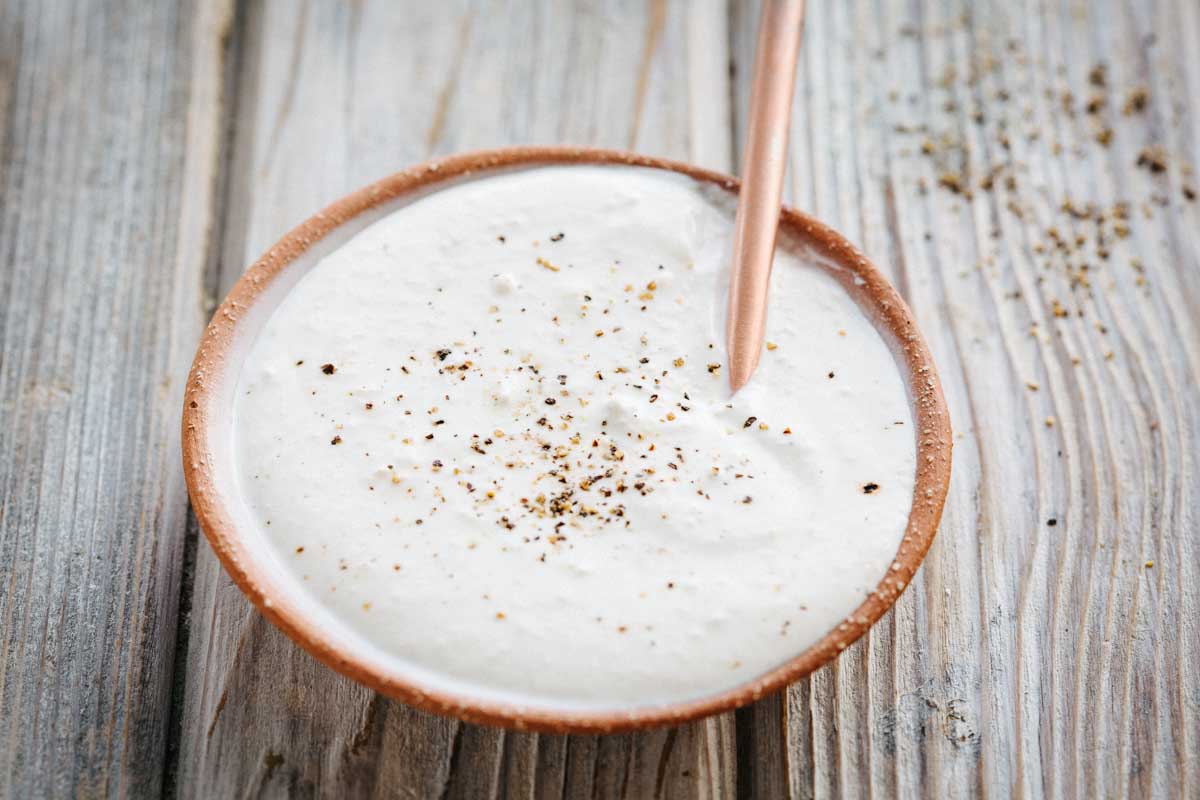 A small terracotta bowl of horseradish sauce on a wooden surface, sprinkled with cracked pepper and a copper spoon inside.