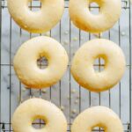 Six pale, freshly glazed Meyer lemon donuts resting on a wire rack with drips of glaze on the surface below.