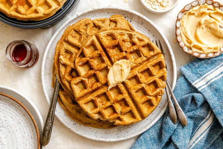 Crispy oatmeal waffles topped with a pat of butter and drizzled with maple syrup on a plate, with a blue linen napkin alongside.