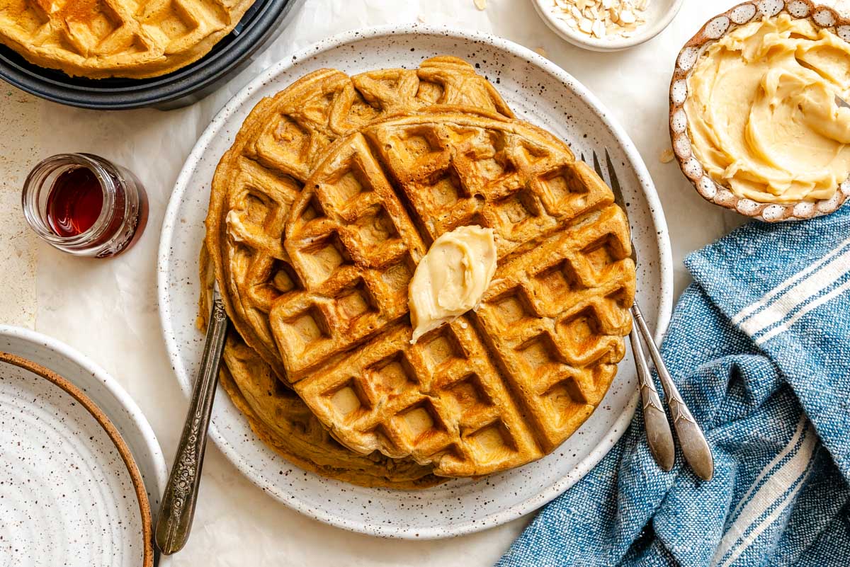 Crispy oatmeal waffles topped with a pat of butter and drizzled with maple syrup on a plate, with a blue linen napkin alongside.
