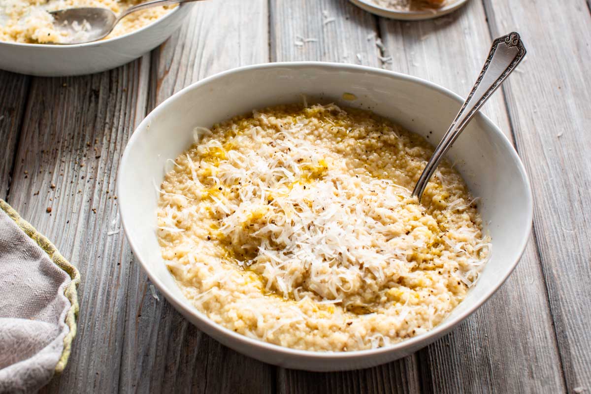 A bowl of creamy pastina topped with grated cheese and pepper, with a spoon dipped in and a block of Parmesan in the background.