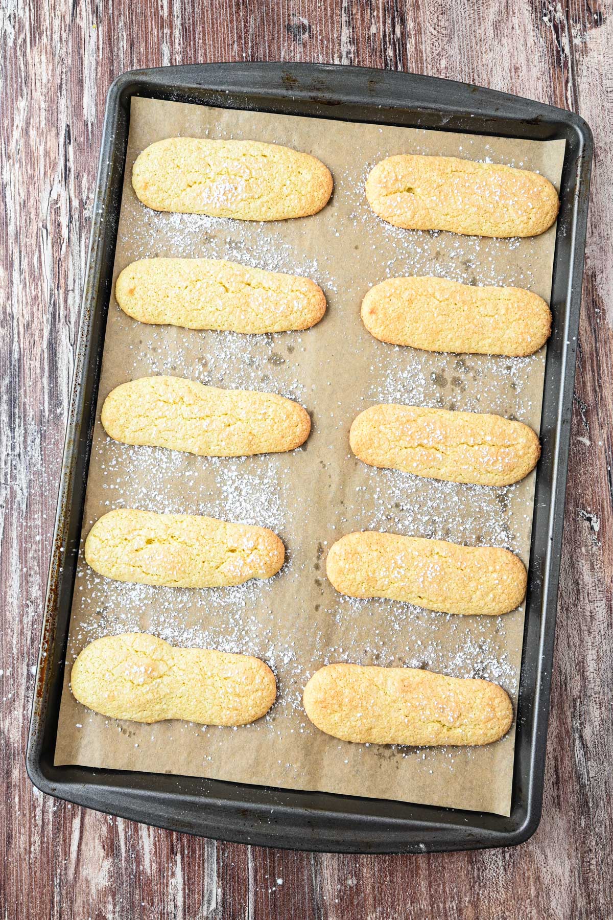 Homemade ladyfingers arranged on a parchment lined baking sheet, lightly dusted with powdered sugar.