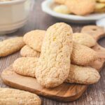 A small stack of crisp, golden ladyfingers displayed on a wooden board with more cookies in the background.