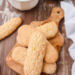Ladyfingers on a wooden board next to a cup of coffee dusted with cinnamon and a plate of additional cookies.