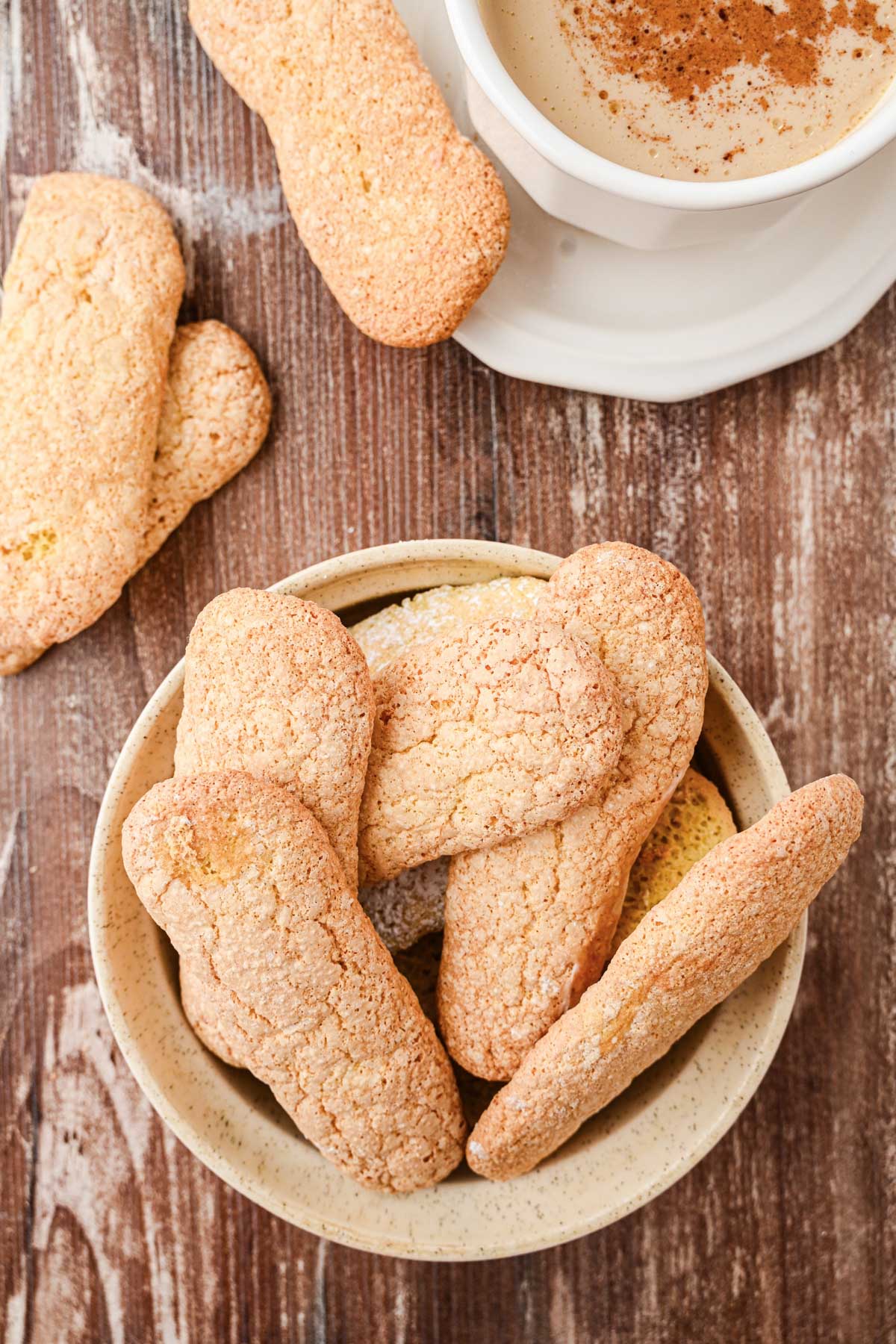 A bowl filled with homemade ladyfingers with a cup of coffee and extra cookies nearby.