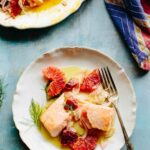 Two plates filled with slow roasted salmon, blood oranges and fennel on a blue surface with a colorful towel alongside.