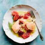 A close up of a plate of roasted salmon with blood orange pieces, thin fennel slices and olive oil on a blue painted background.