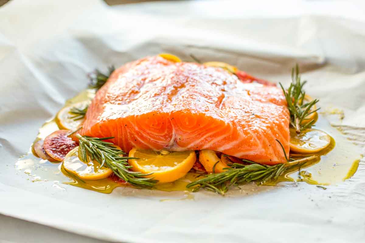 A raw salmon fillet set over lemon and orange slices with rosemary and olive oil on parchment paper ready for slow roasting.