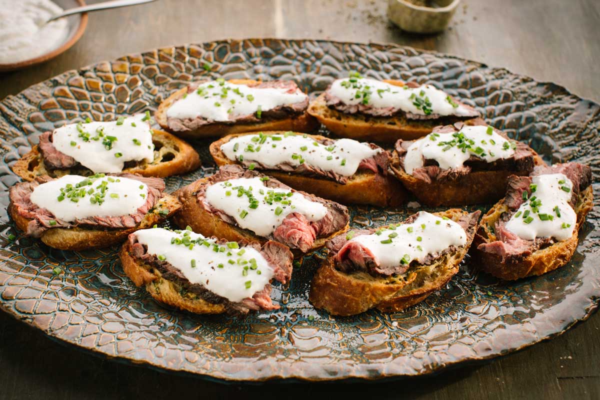 A large textured platter filled with toasted baguette slices topped with thin sliced steak, horseradish sauce and chopped chives.