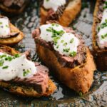 A group of steak crostini arranged on a patterned serving plate, each topped with white horseradish sauce and chopped chives.