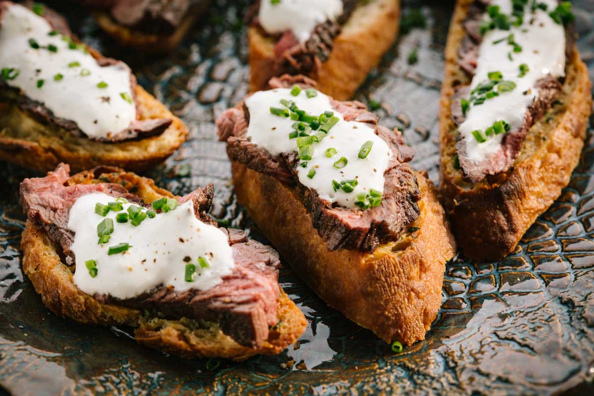 A group of steak crostini arranged on a patterned serving plate, each topped with white horseradish sauce and chopped chives.