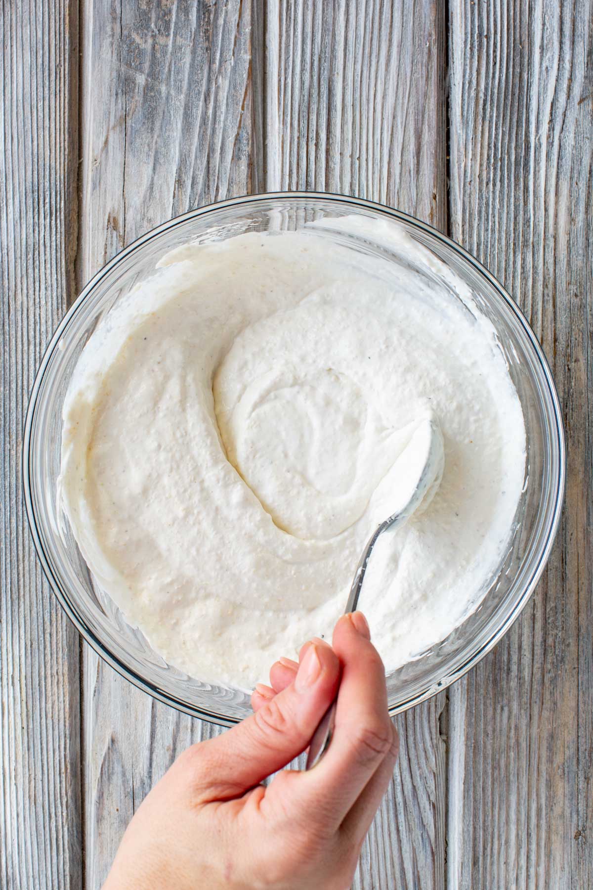A hand holds a spoon swirling creamy white horseradish sauce in a glass bowl on a weathered wooden surface.