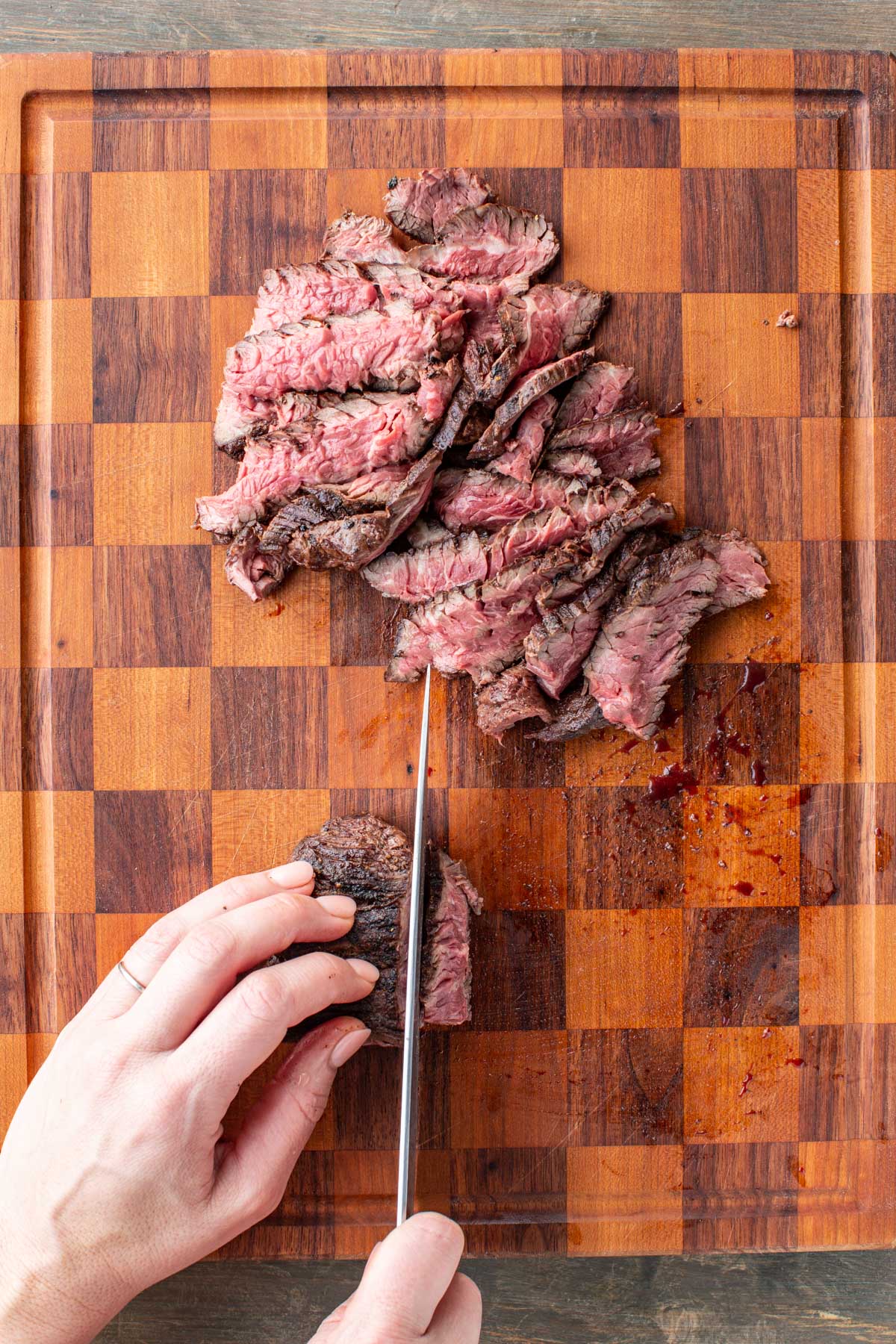 Hands use a sharp knife to cut thin slices of medium rare steak on a checkered wooden cutting board.