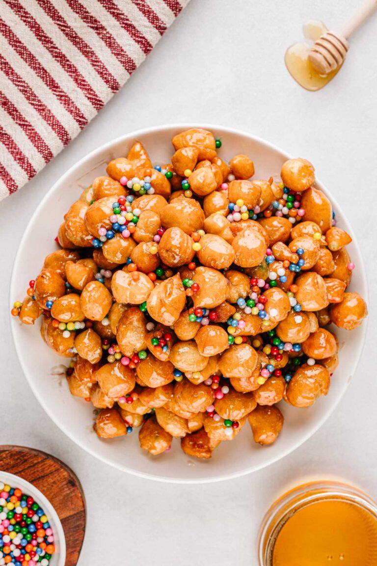 A mound of honey coated struffoli piled high on a white plate and topped with colorful sprinkles.