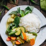 Plate of Thai green vegetable curry served alongside jasmine rice and garnished with Thai basil leaves.