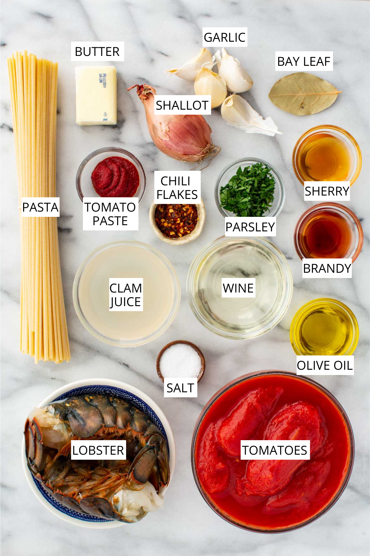 Overhead layout of lobster fra diavolo ingredients including lobster, pasta, tomatoes, tomato paste, garlic, shallot, chili flakes, parsley, bay leaf, butter, olive oil, wine, sherry, brandy, clam juice, and salt on a marble surface.