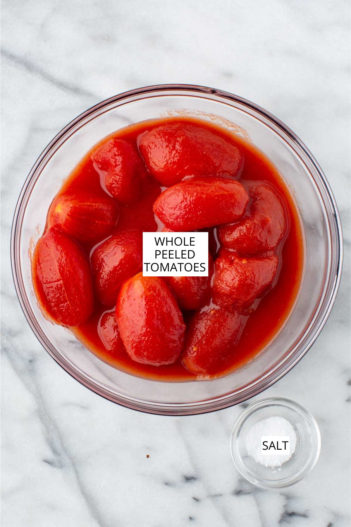 Whole peeled tomatoes in their juices in a glass bowl with a small dish of salt on a marble surface.