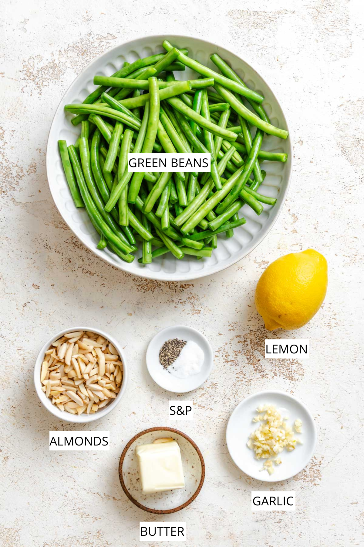 Overhead layout of ingredients for green beans amandine including green beans, lemon, sliced almonds, butter, garlic, salt, and pepper labeled on a light surface.