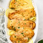 Chicken francaise arranged on an oval white platter with lemon slices, spooned with lemon butter sauce and finished with chopped parsley.