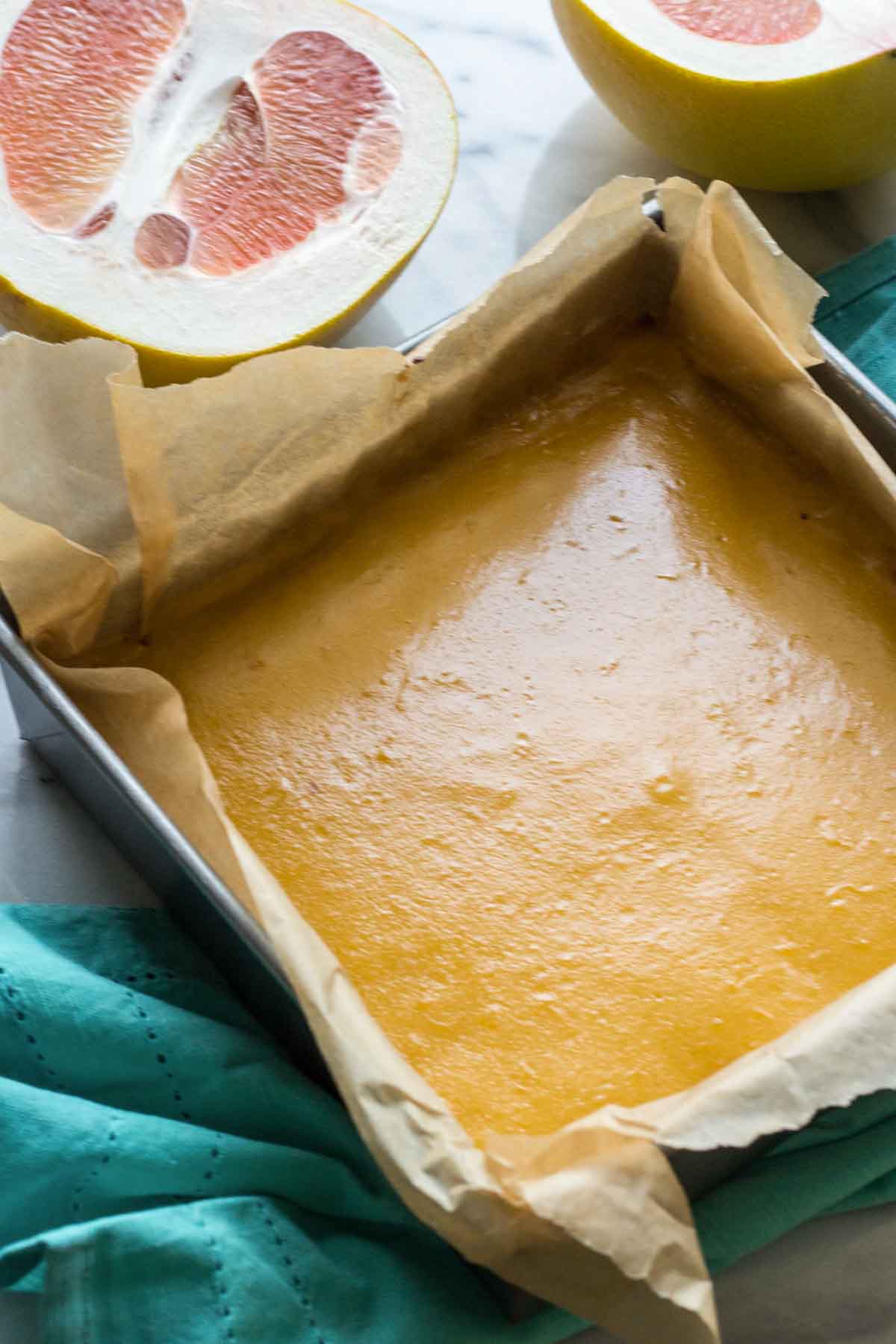 Fresh grapefruit curd poured into a parchment-lined square baking pan, with halved grapefruits in the background.