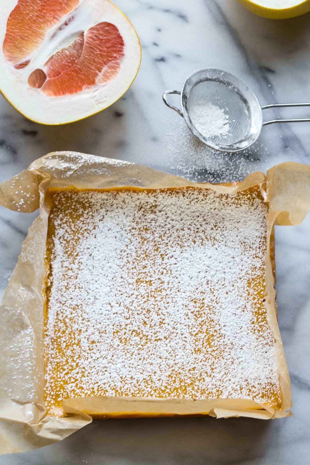 Overhead view of grapefruit bars fully dusted with powdered sugar in a parchment-lined pan, with a grapefruit half nearby.