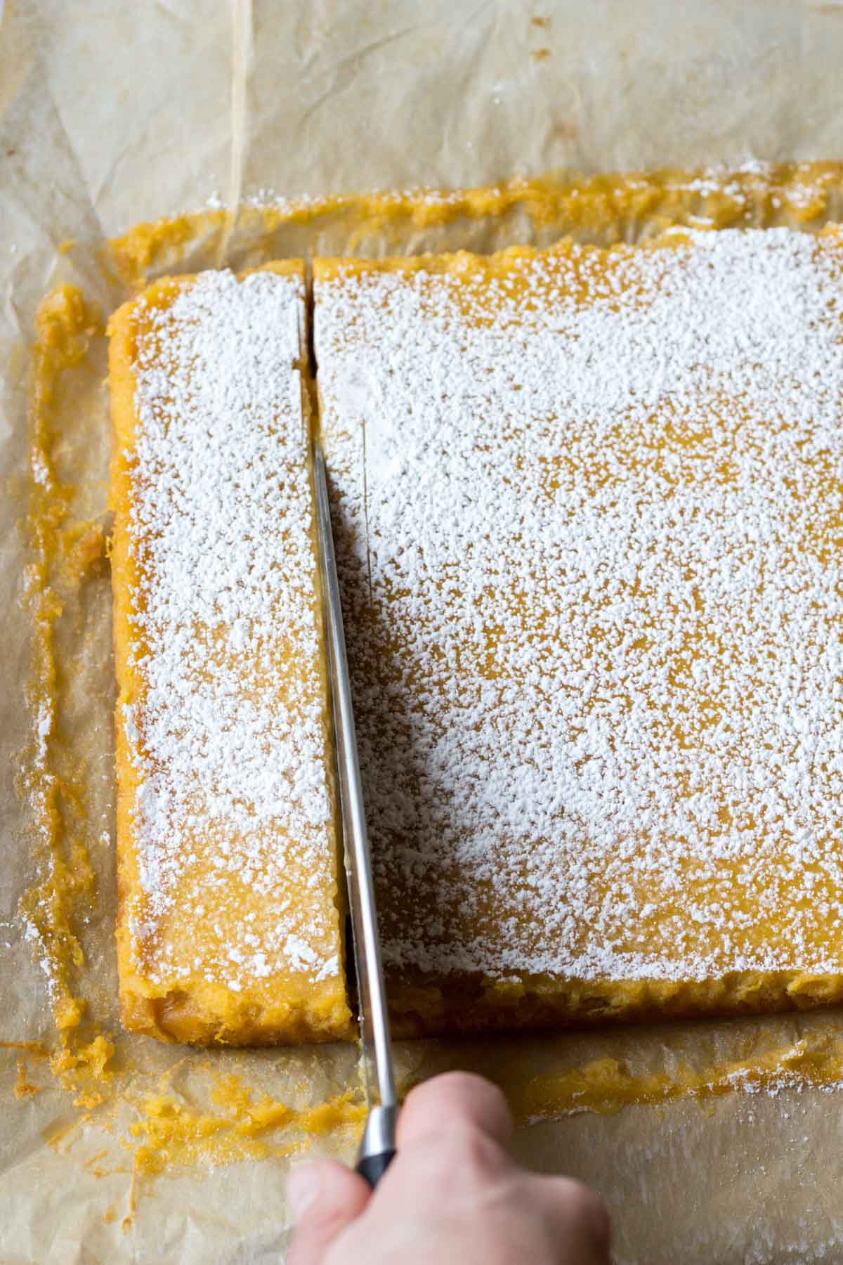 A sharp knife slicing powdered sugar-topped grapefruit bars into neat squares in the pan.