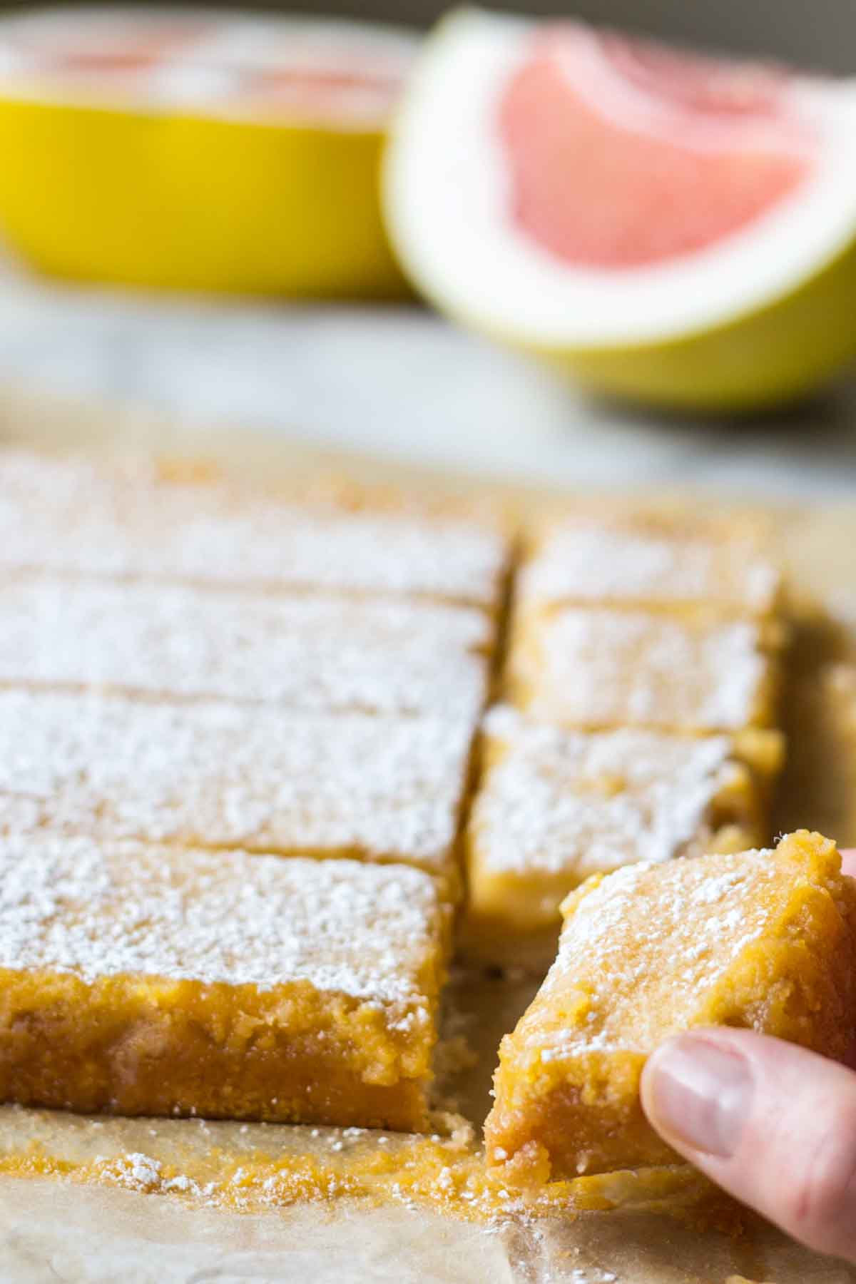 A hand lifting a single grapefruit bar from the pan, showing the creamy curd and sturdy crust.