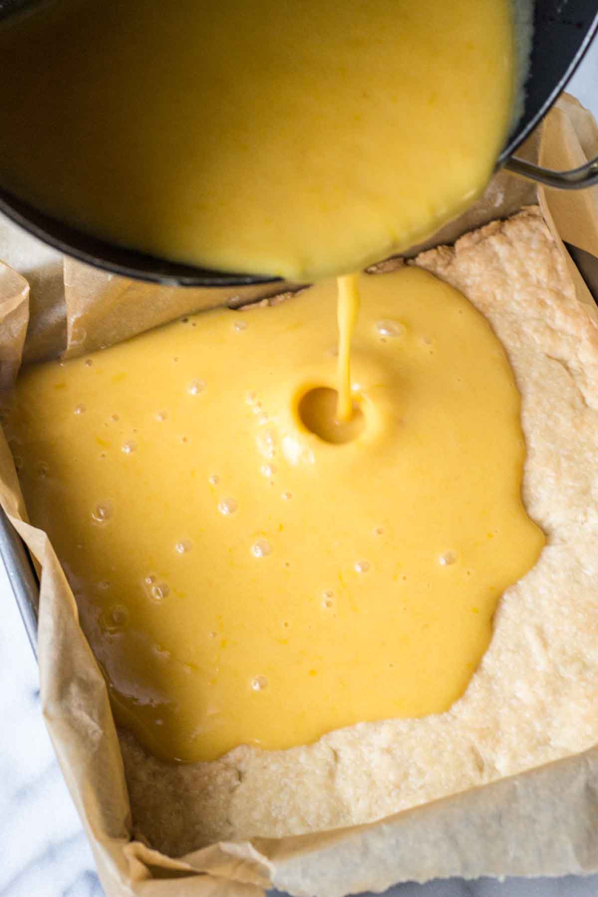 Warm grapefruit curd being poured from a saucepan onto a baked shortbread crust in a parchment-lined pan.