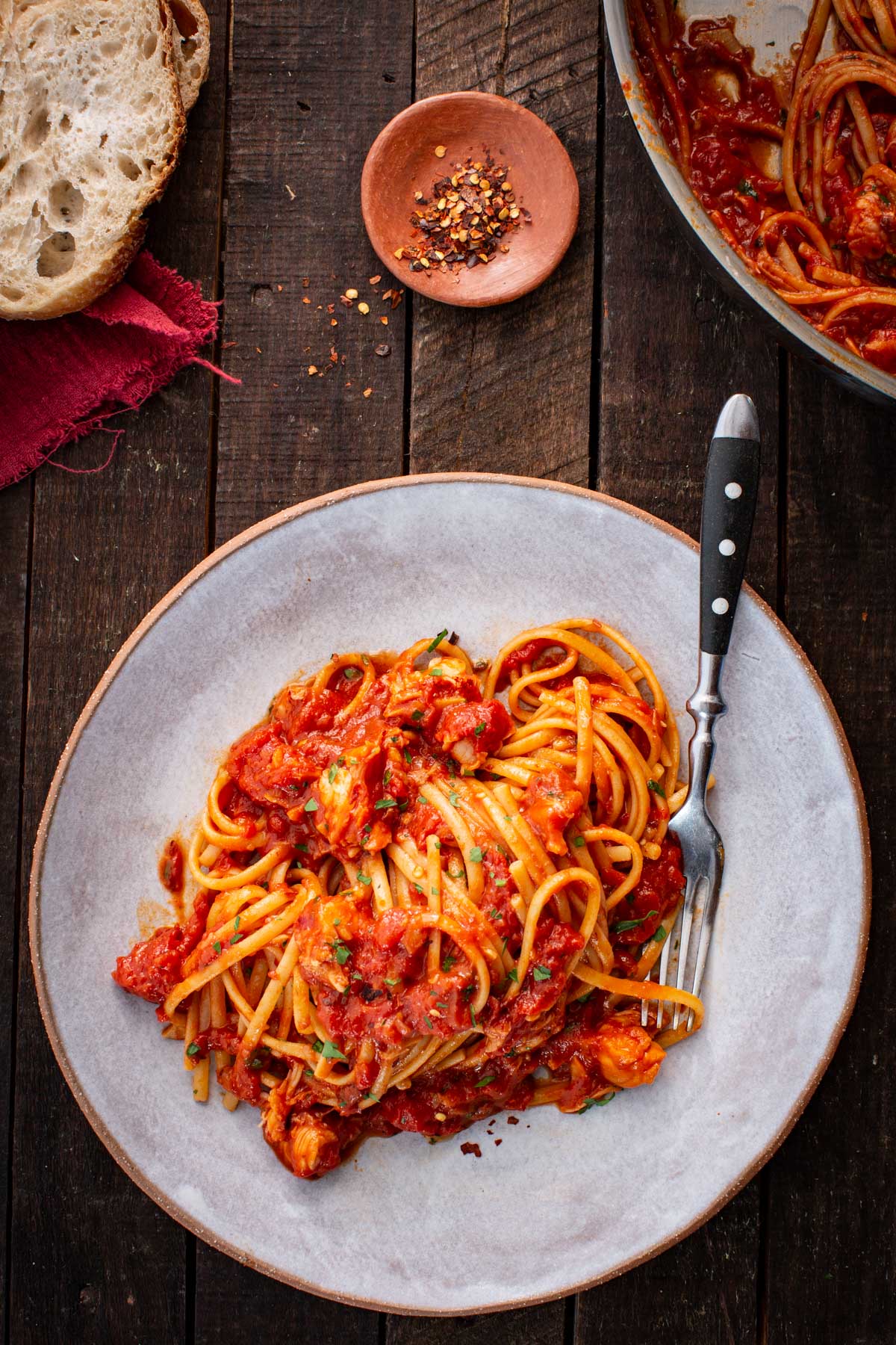 Plated serving of lobster fra diavolo with pasta on a shallow gray bowl, set on a dark wooden table with chili flakes and crusty bread nearby.