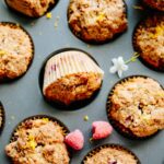 Overhead view of orange raspberry crumb muffins in a muffin tin with golden crumb topping, fresh raspberries and orange zest scattered throughout the scene.