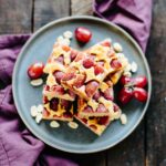 Peanut butter grape shortbread bars stacked on a gray plate with fresh red grapes and sliced peanuts on a wooden surface.