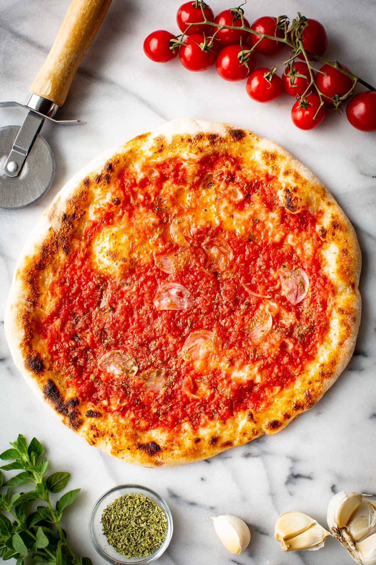 Neapolitan pizza marinara on a marble surface next to cherry tomatoes on the vine, garlic, oregano and a pizza cutter.