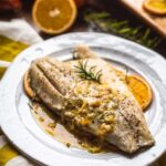 Roasted fish fillet served on a white plate with orange slices and rosemary, spooned with pan juices and citrusy sauce.
