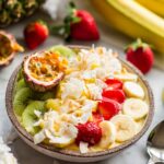 A tropical smoothie bowl surrounded by fresh fruit ingredients including bananas, pineapple, strawberries, kiwi, and passion fruit on a marble surface.