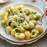 Side view of hands eating a plate of broccoli pasta with a fork.