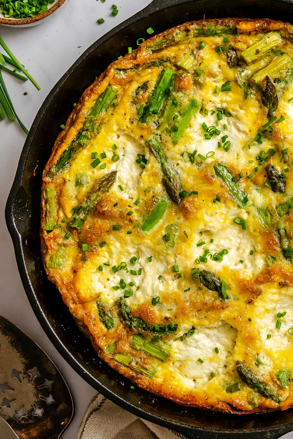 Close up of a golden asparagus ricotta frittata in a cast iron skillet with tender asparagus pieces, creamy ricotta and fresh chives sprinkled on top.