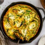 Overhead view of a cast iron skillet with asparagus ricotta frittata as a slice is lifted out, revealing the fluffy egg interior with asparagus and ricotta.