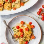 Overhead shot of chicken scallopini served on a large white oval platter with a spoon, alongside an individual portion plated on a round gray dish with a fork, set on a marble surface with fresh cherry tomatoes on the vine and basil leaves.