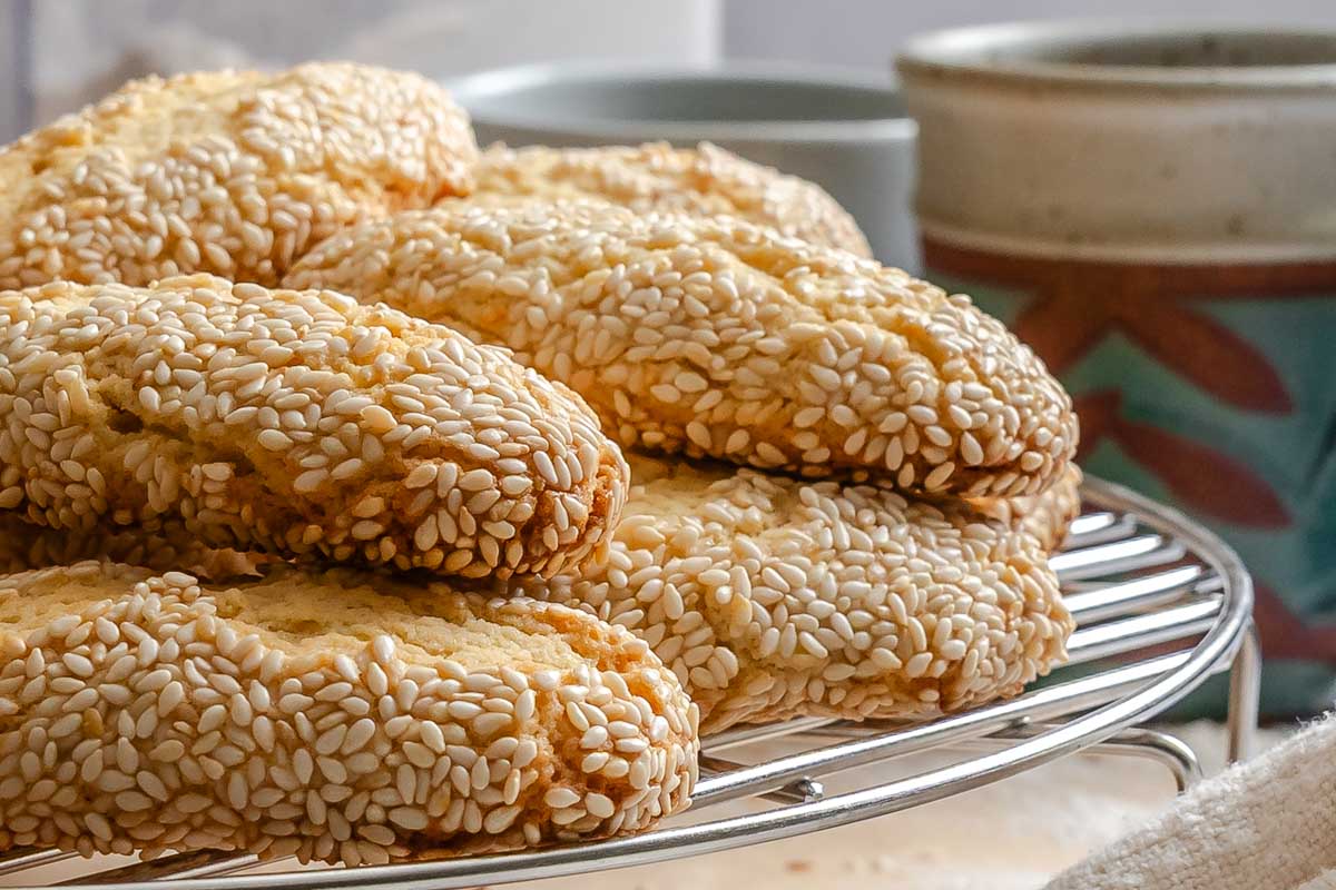 Side view of Italian sesame seed cookies piled high on a wire cooling rack, with two ceramic mugs blurred in the background and a linen cloth in the foreground.